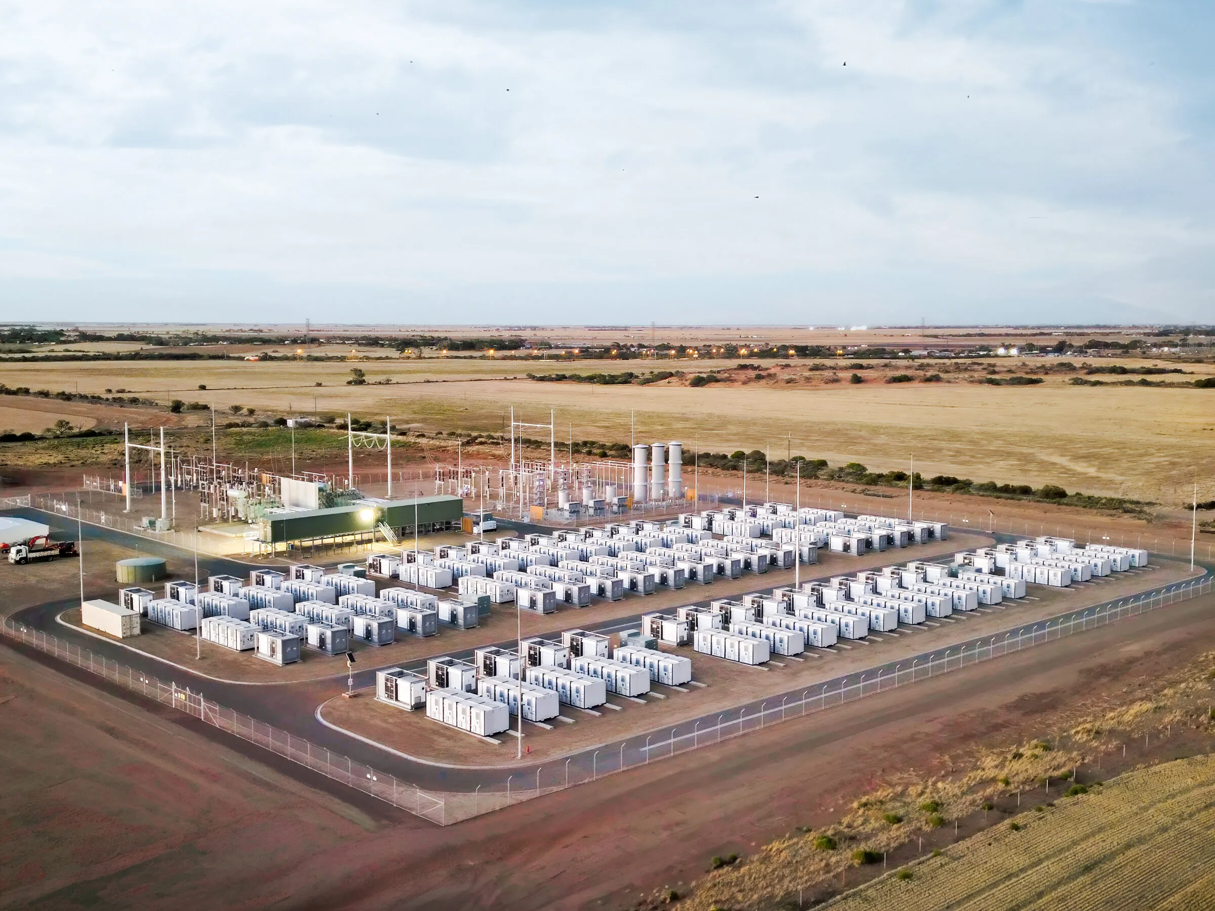 Aerial view of a battery energy storage site on flat open country, dozens of white enclosures in ranks beside a substation and switchyard