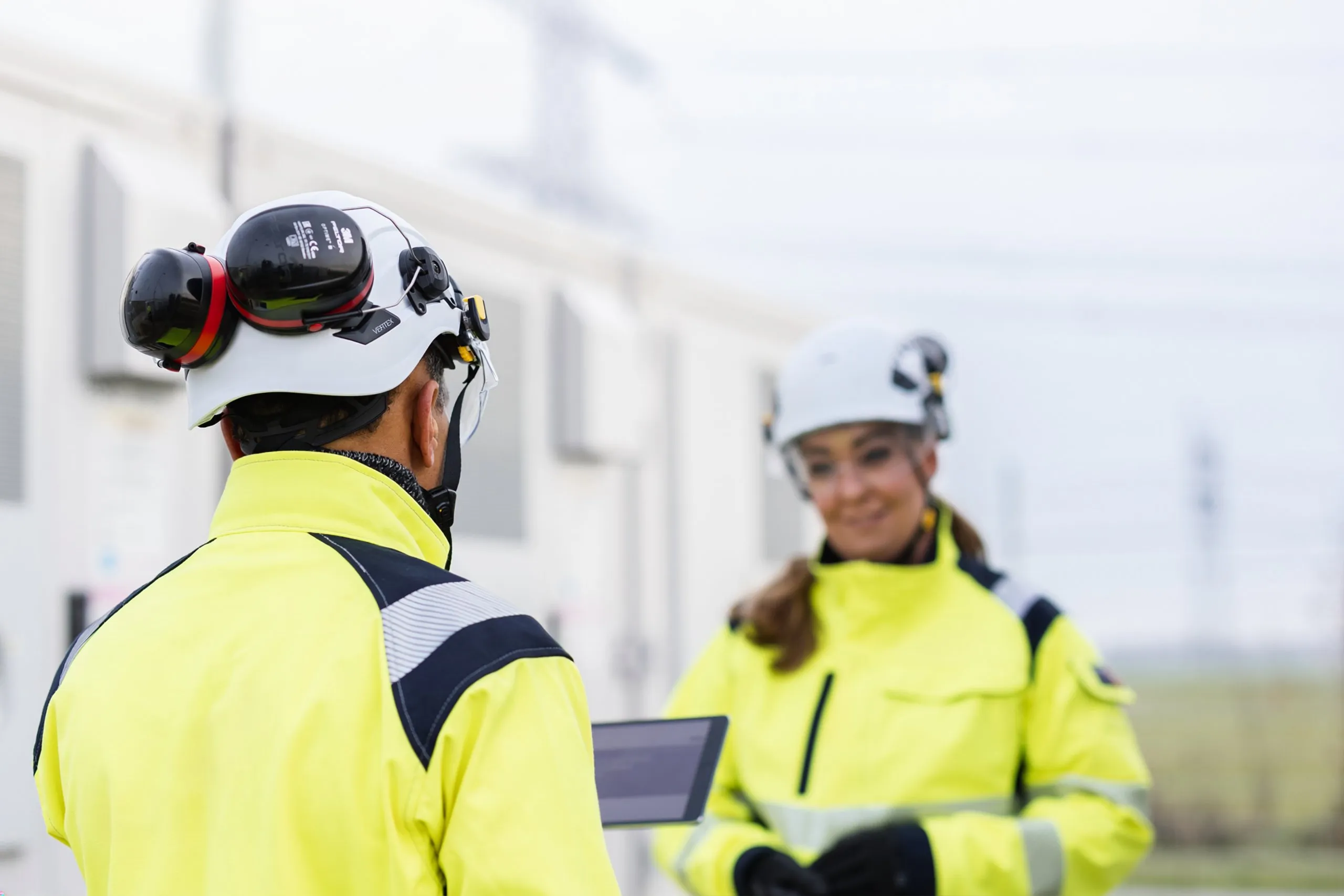 Two technicians in high-visibility gear and hard hats, one holding a tablet, facing each other.
